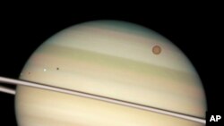 FILE - This image provided by NASA captures the transit of several moons across the face of Saturn. The giant orange moon Titan – larger than the planet Mercury – can be seen at upper right.